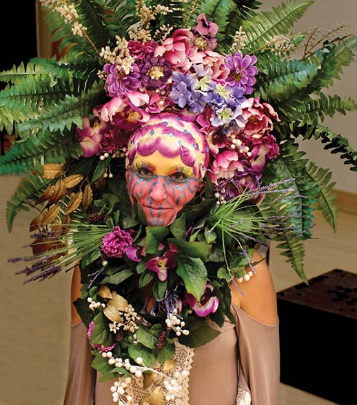 An actress poses in her stage attire: a headdress made out of flowers. Her face is painted to look like a flower.