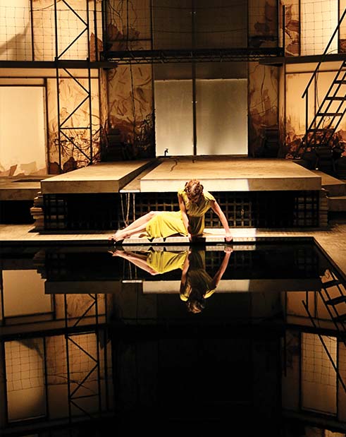An actress in yellow sits above a mirror, looking down at herself while performing.